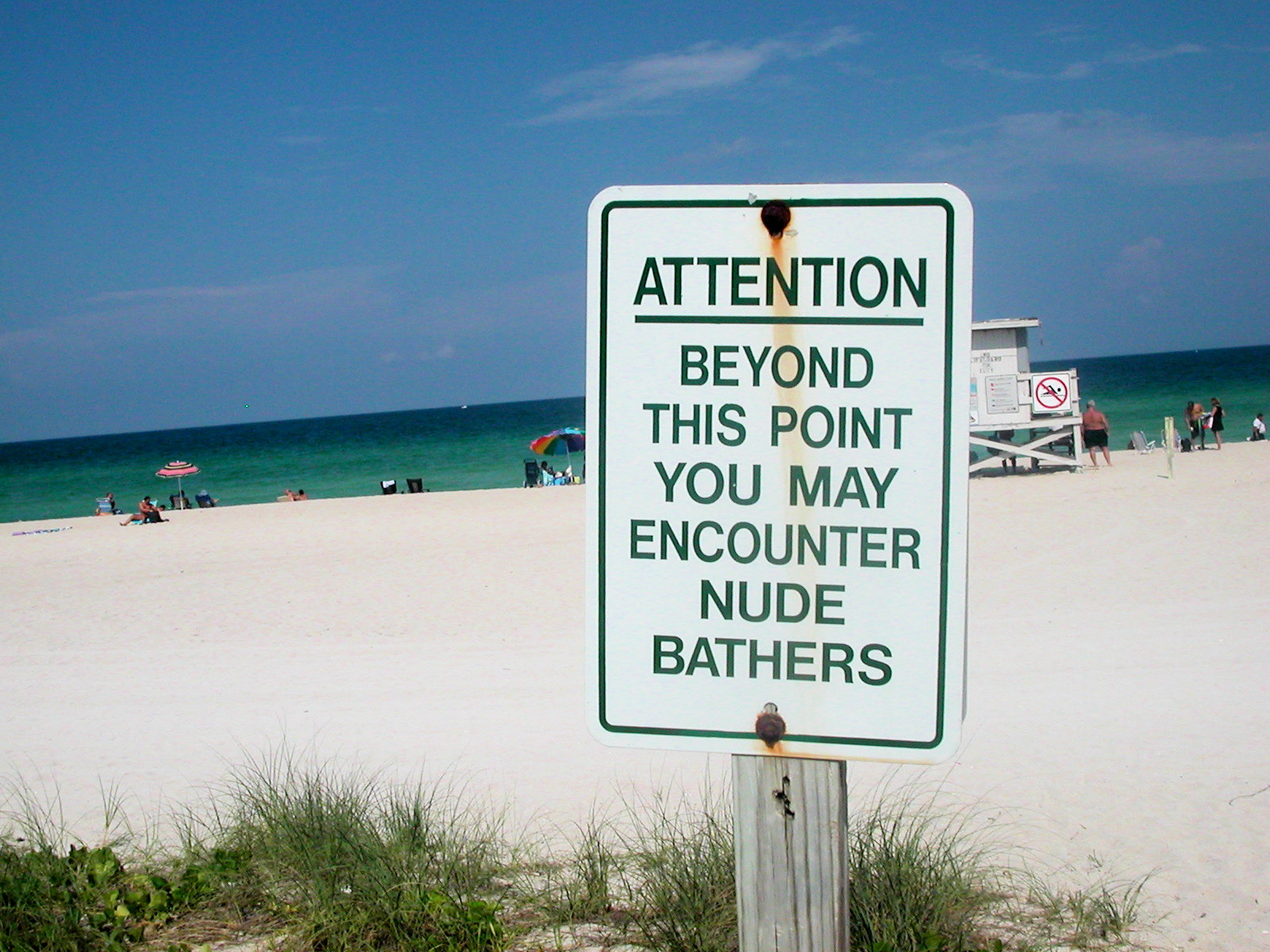 Photo of a slightly rusted sign in the foreground with a beach in the background. The sign has green letters on a white backdrop and it reads, "Attention: Beyond this point you may encounter nude bathers."