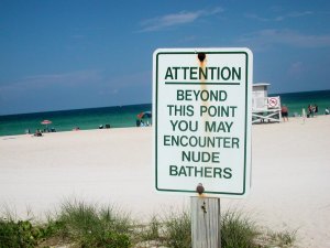 Photo of a slightly rusted sign in the foreground with a beach in the background. The sign has green letters on a white backdrop and it reads, "Attention: Beyond this point you may encounter nude bathers."