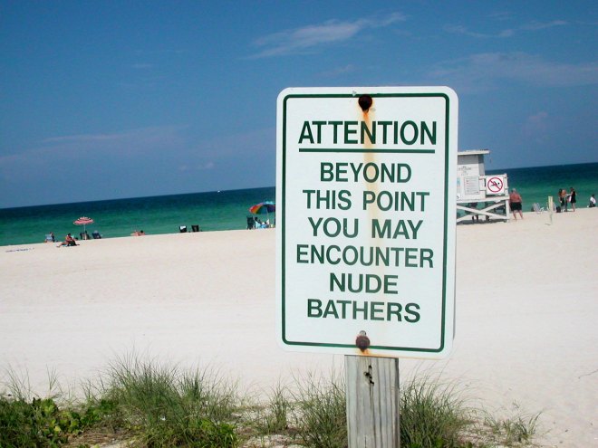 Photo of a slightly rusted sign in the foreground with a beach in the background. The sign has green letters on a white backdrop and it reads, "Attention: Beyond this point you may encounter nude bathers."