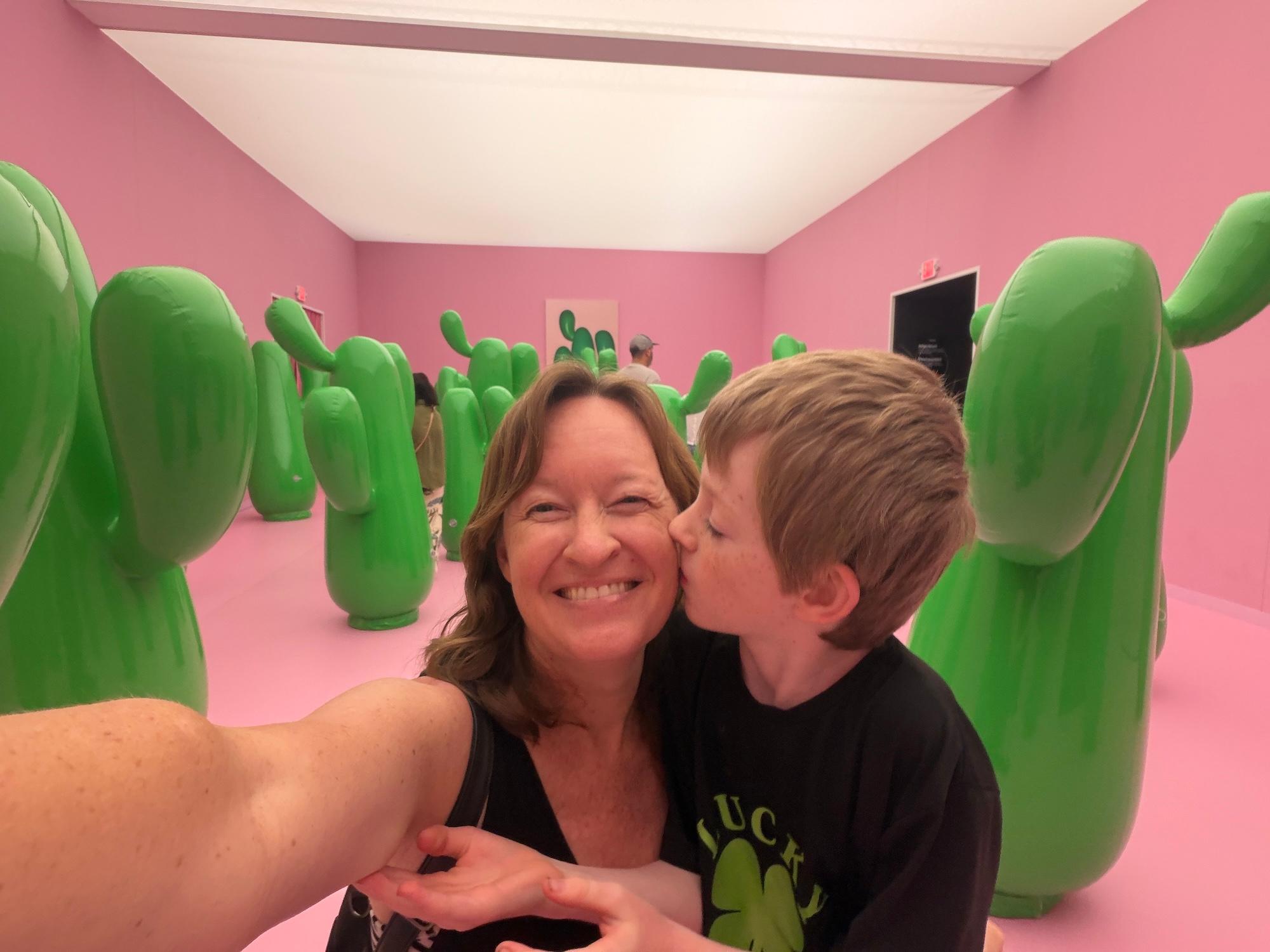 photo of writer Ciara LaVelle taking a selfie with her son giing her a kiss on the cheek inside an immersive museum with cactus sculptures in the background