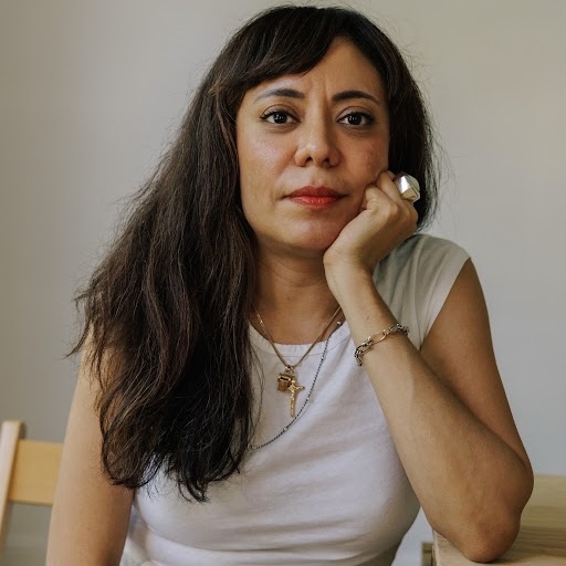 headshot of a young woman with dark hair wearing a sleeveless white top, seated in a wooden chair and resting her chin on her left hand with her elbow leaning on a wooden table