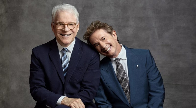 Portrait of comedians Steve Martin and Martin Short in suits. Short leans his head on Martin's shoulder.