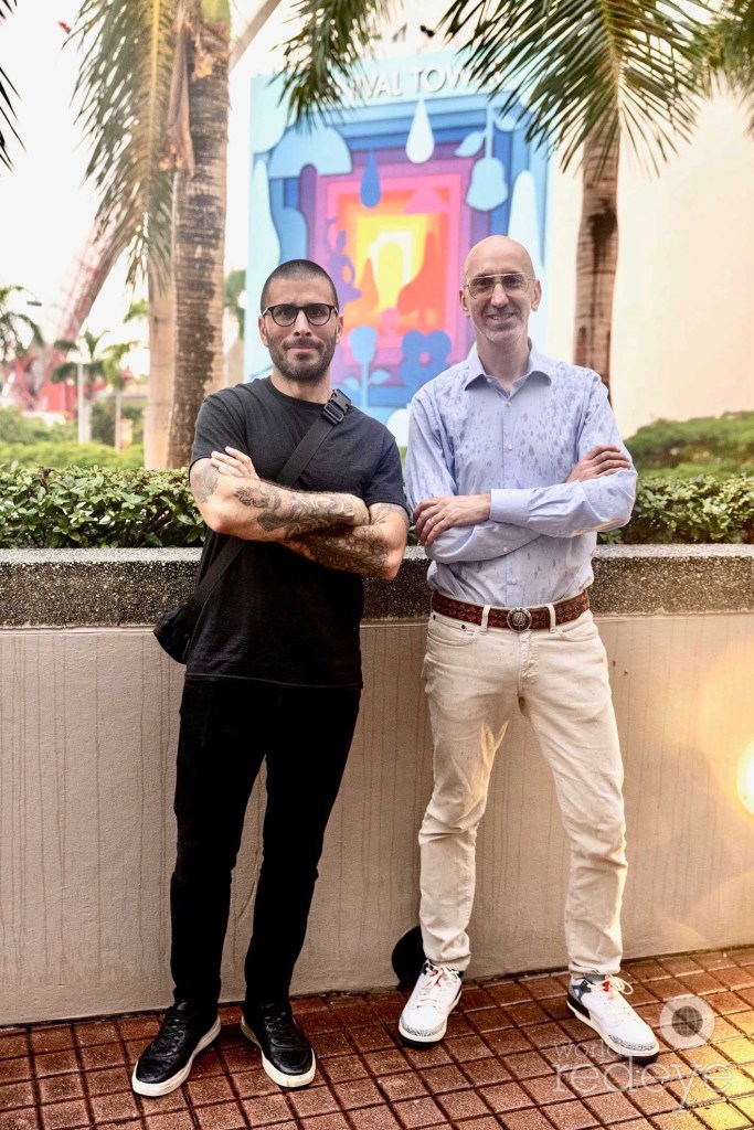 two men pose in front of a mural in the distance