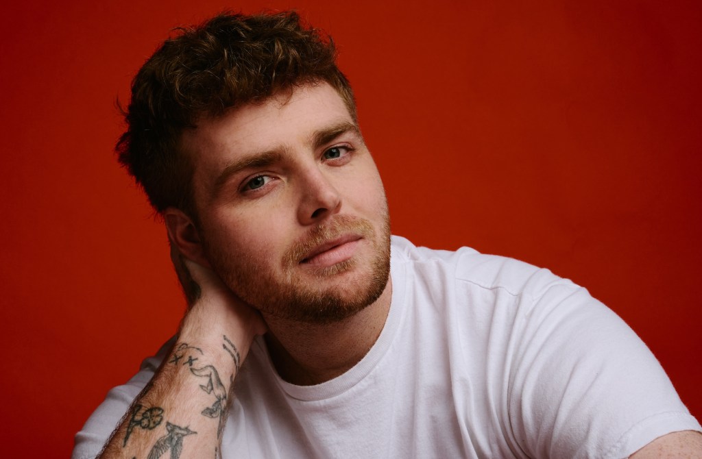 portrait of singer Alex Warren in a white tee posing in front of a red background