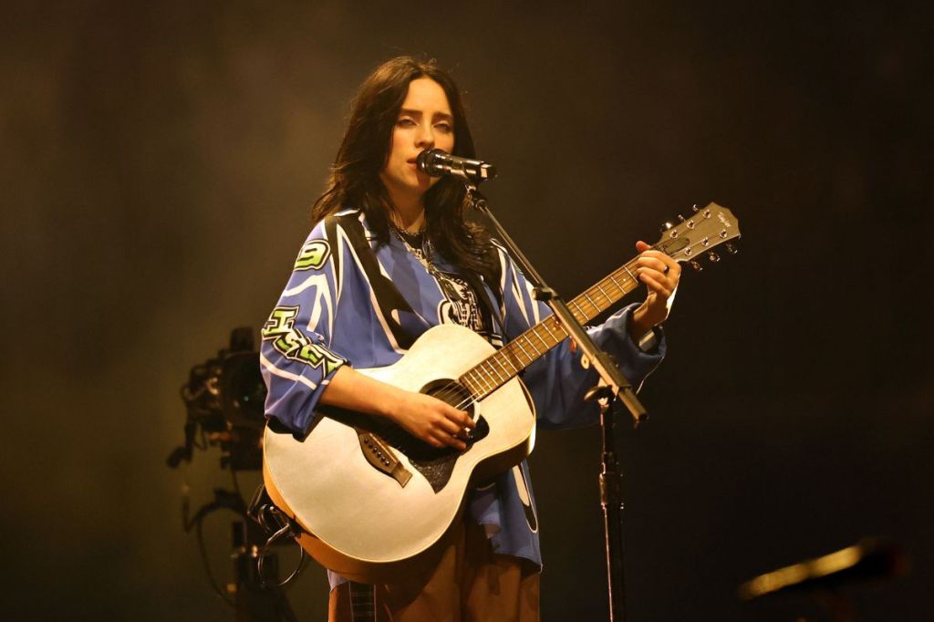 Billie Eilish playing an acoustic guitar on stage.