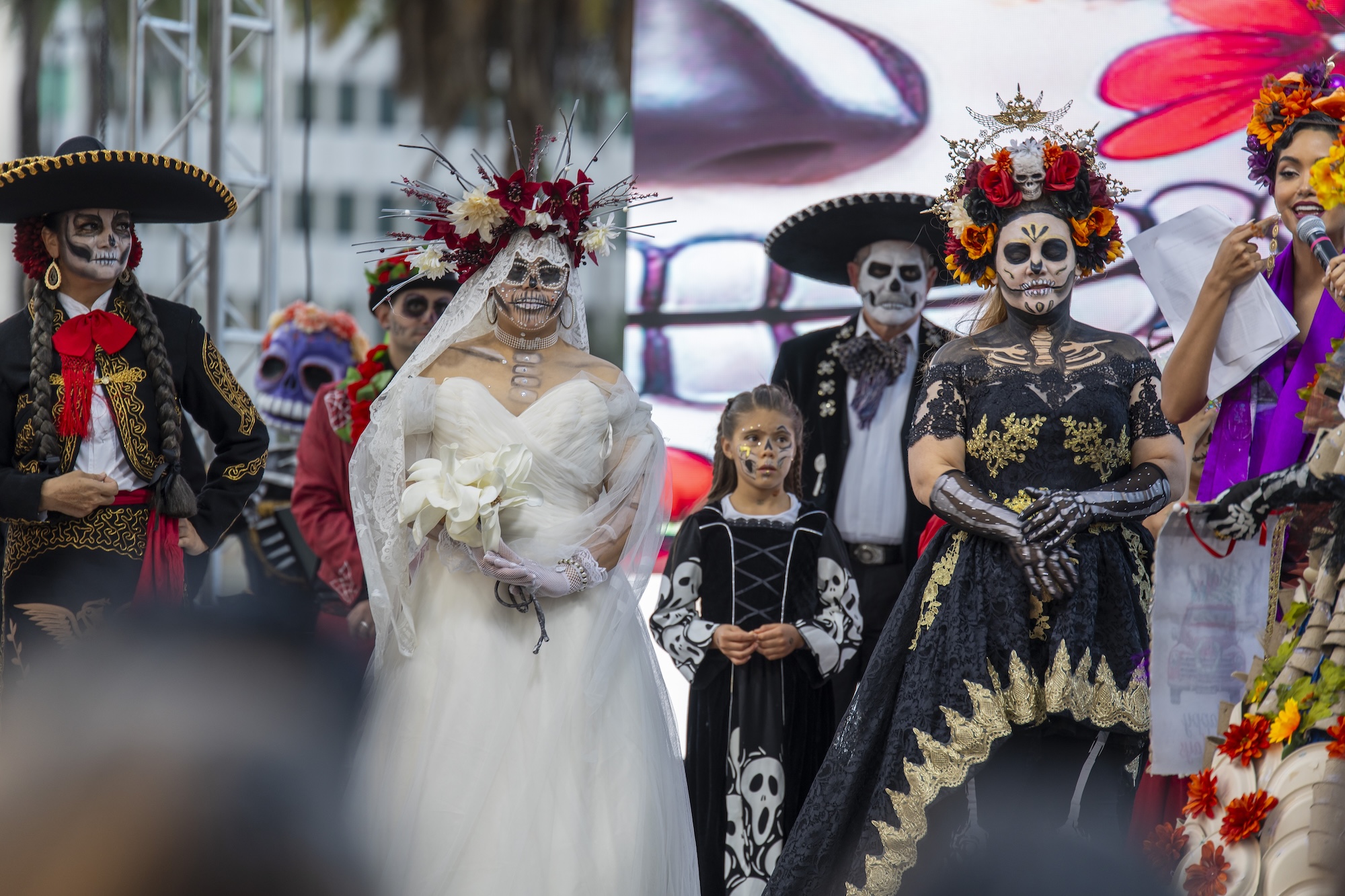 six adults and a young girl dressed in Day of the Dead costumes. All have skeletal makeup on. One has a wedding dress on; others wear mariachi costumes