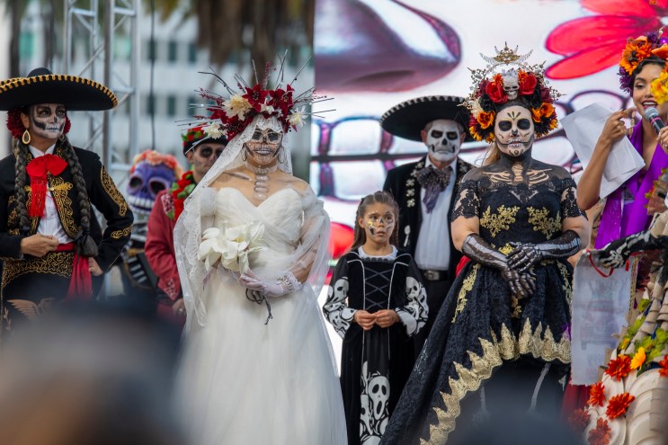 six adults and a young girl dressed in Day of the Dead costumes. All have skeletal makeup on. One has a wedding dress on; others wear mariachi costumes