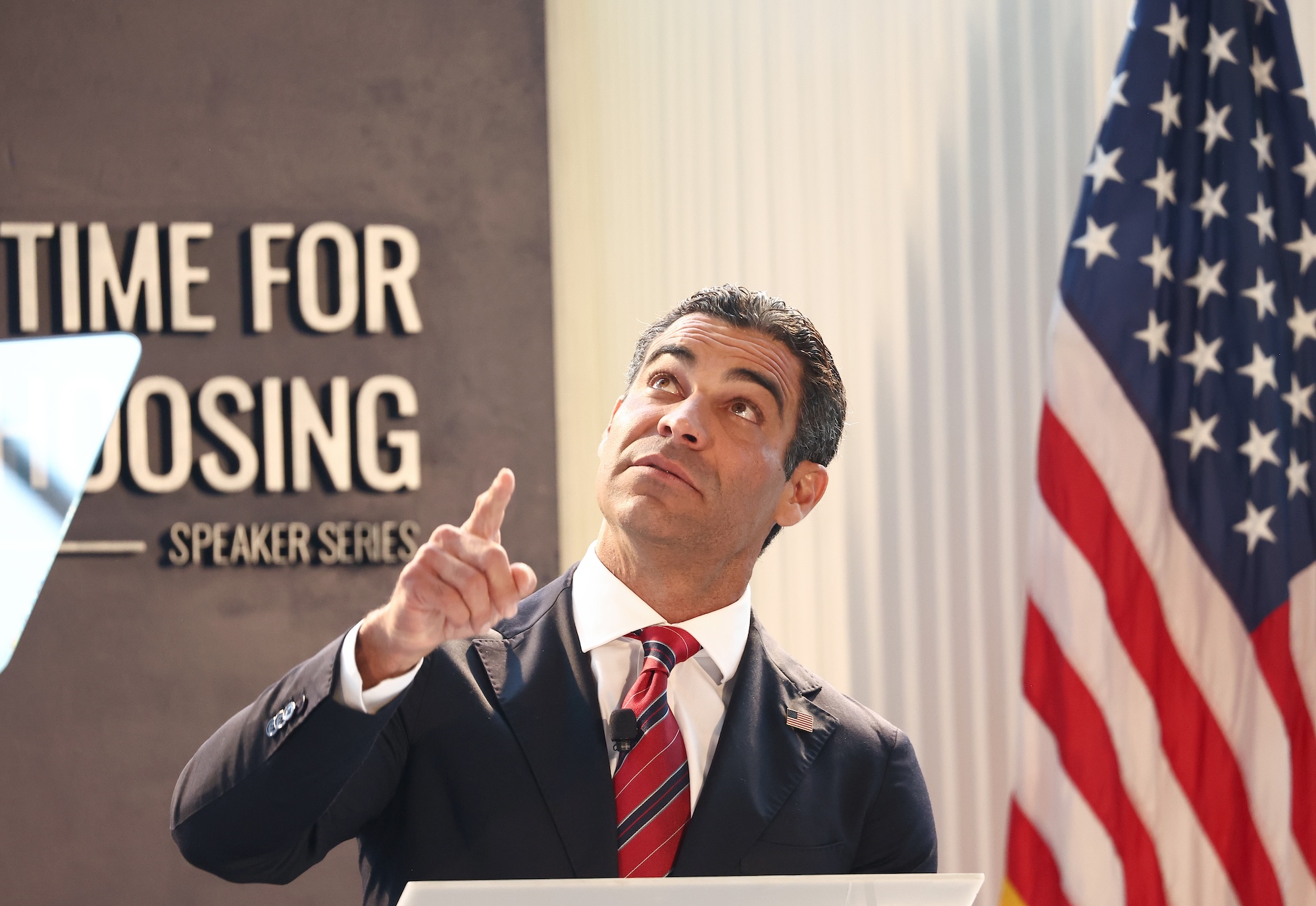 photo of a man wearing a suit and tie, speaking at a podium at the Ronald Reagan Presidential Library in Simi Valley, California, while gazing and pointing vaguely upward. An American flag is in the background.