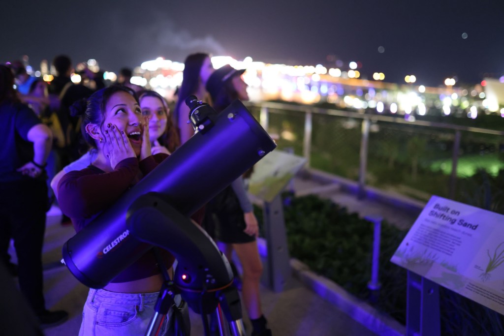 a woman looking up at the sky next to a telescope looks surprised