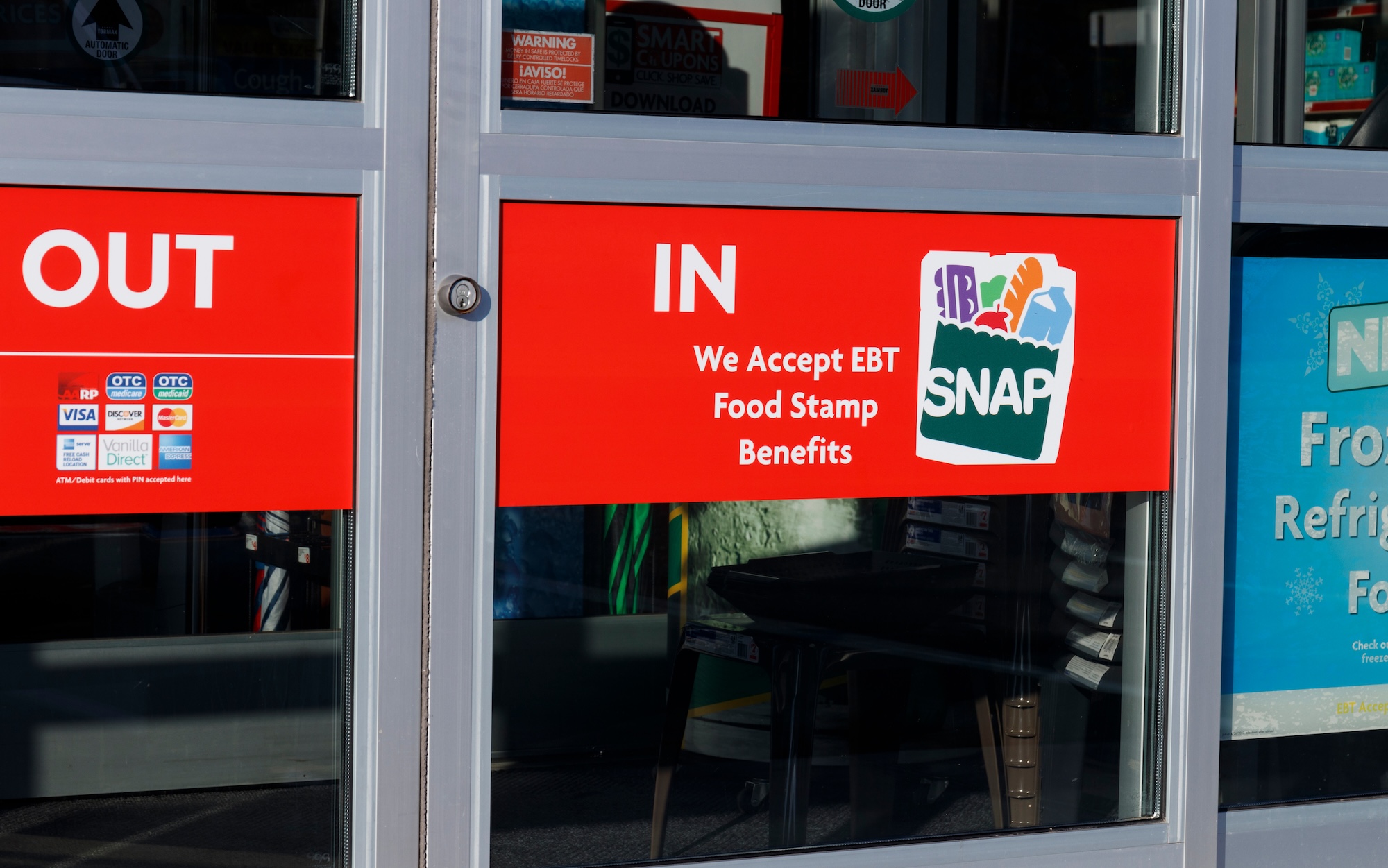 close-up photo of a store entrance with a sign that reads "We Accept EBT Food Stamp Benefits" and the SNAP logo, which shows the acronym on an illustration of grocery bag containing a milk carton, a loaf of french bread, an apple, a green vegetable, and a carton of eggs