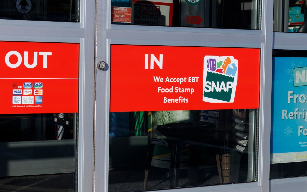 close-up photo of a store entrance with a sign that reads "We Accept EBT Food Stamp Benefits" and the SNAP logo, which shows the acronym on an illustration of grocery bag containing a milk carton, a loaf of french bread, an apple, a green vegetable, and a carton of eggs