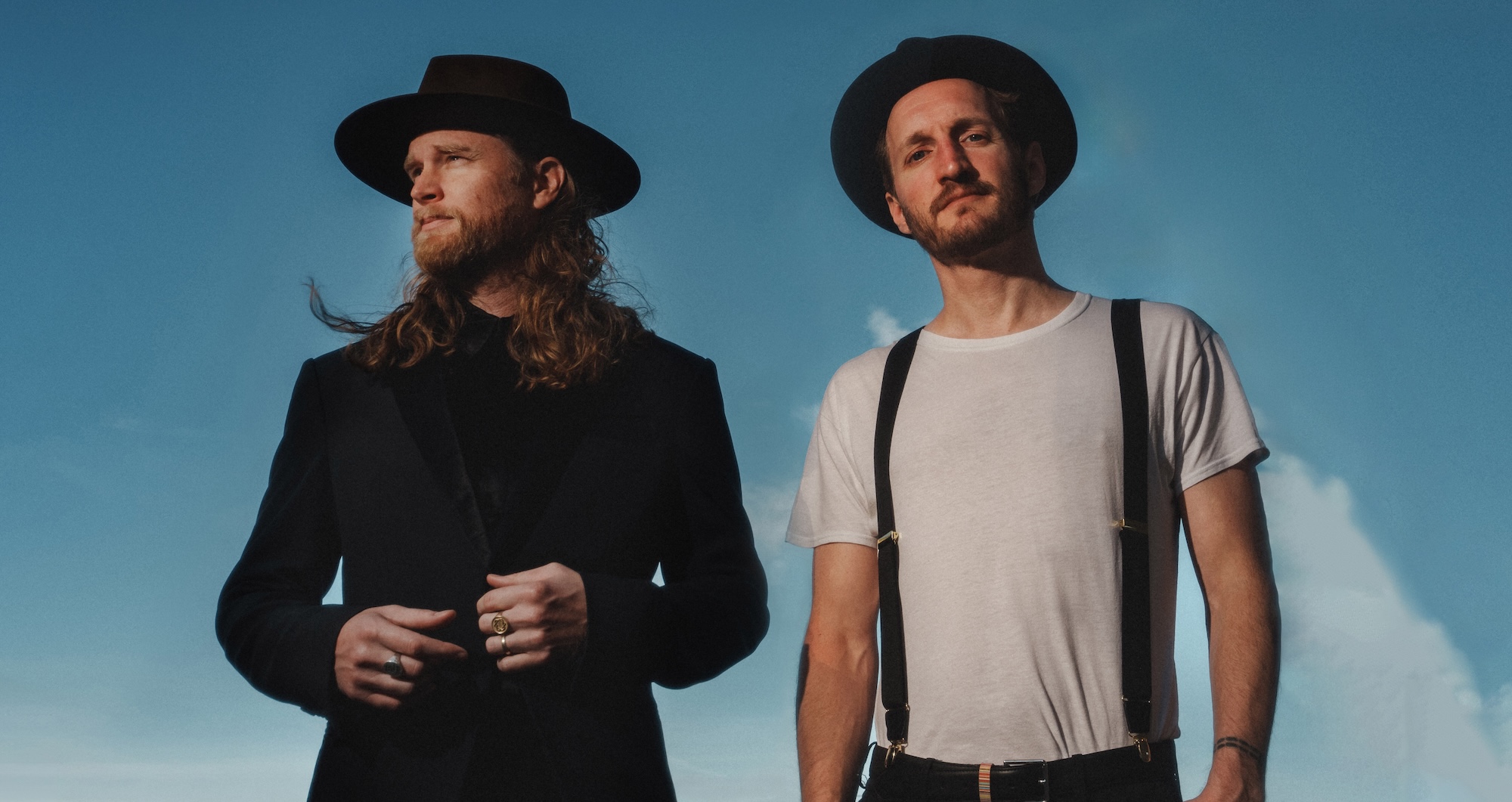 two men in fedoras pose with the sky in the background