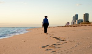 photo of a fully-clothed woman walking along the shore on the beach with high-rise buildings in the background on the right