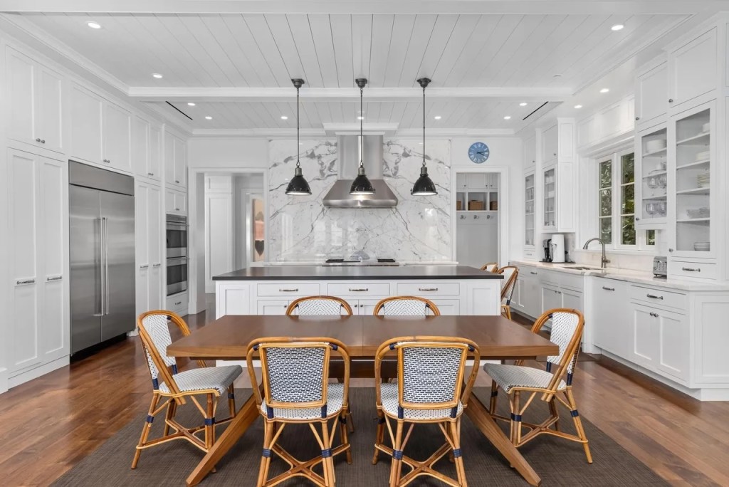 a wooden dining table sits in the foreground of a kitchen accented with stainless steel appliances and a wall of marble