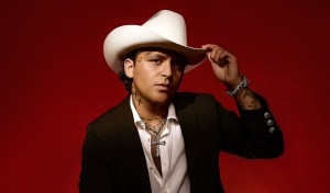 photo of singer Christian Nodal wearing a cream cowboy hat and posing against a red backdrop