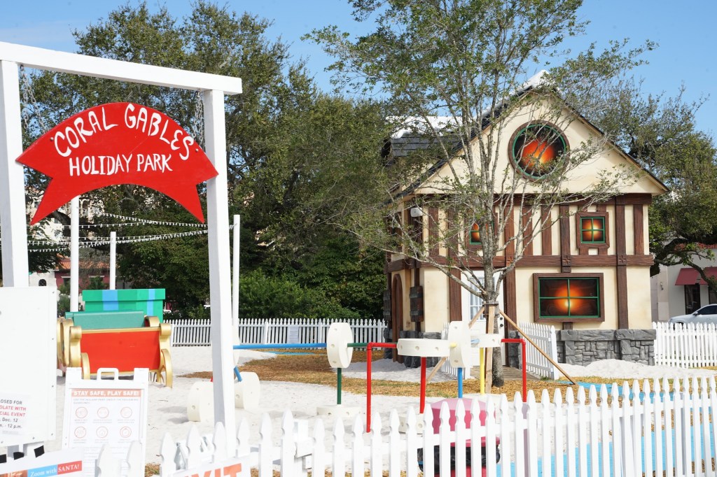 photo of a holiday village with fake snow on the ground, a playground, a house, and a wooden red sign on white wooden poles reading "Coral Gables Holiday Park"