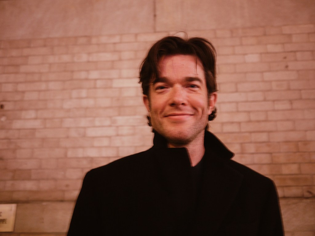portrait of comedian John Mulaney in a black coat, smiling in front of a brick wall