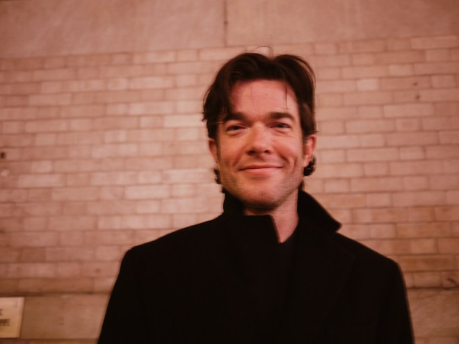 portrait of comedian John Mulaney in a black coat, smiling in front of a brick wall