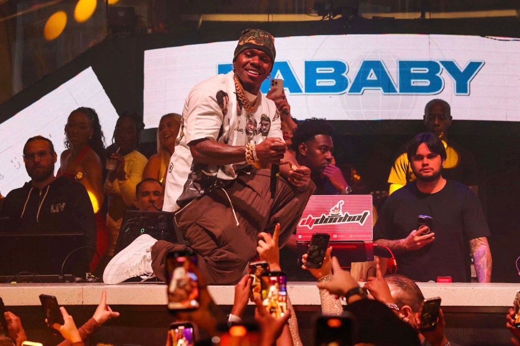 photo of rapper DaBaby kneeling on a DJ booth as he performs in front of   an audience at a club
