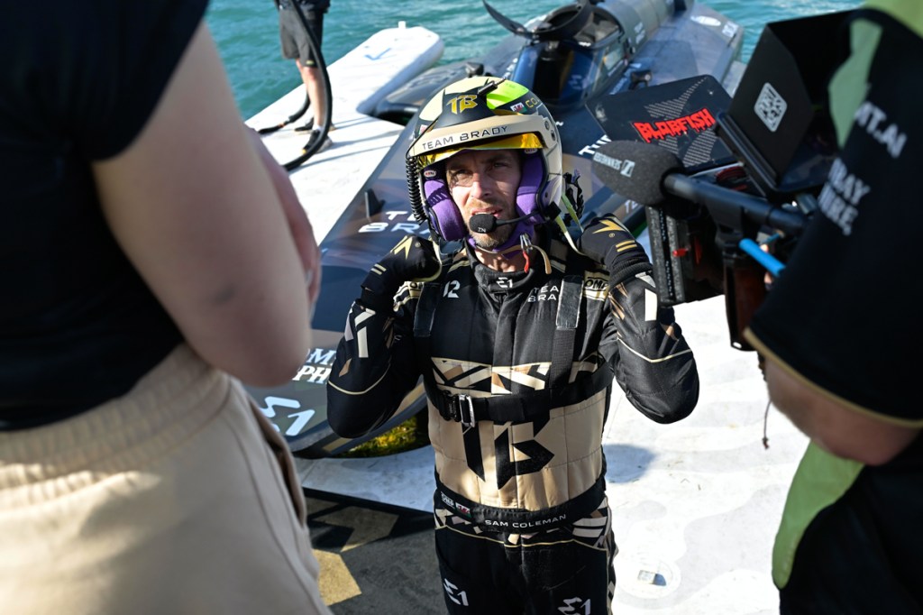 photo of a man in a racing suit and helmet coming out of a boat as a person holding a video camera records him