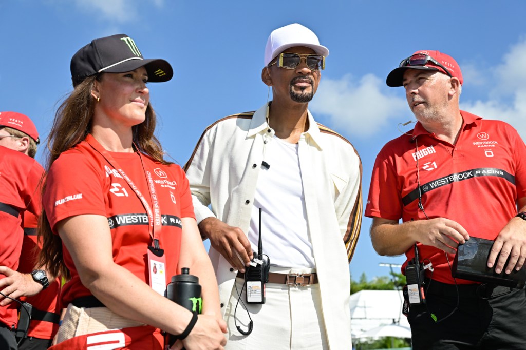 photo of Will Smith in a white baseball cap and pastel pants,shirt, and button-down standing between a man and woman in red shirts and baseball caps