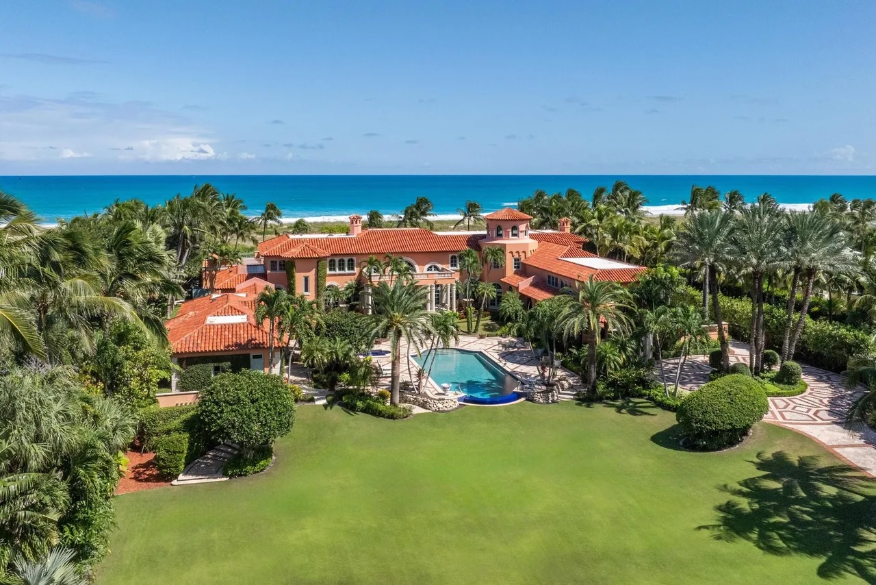 a sprawling Mediterranean-style mansion is surrounded by trees and shrubs with Palm Beach's stark-blue waters in the background