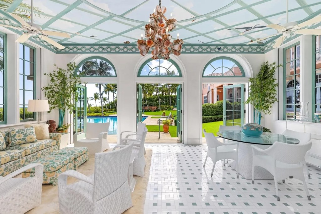 a white-and-mint-colored pool house with a floral chandelier and couch sits just beyond the backyard pool
