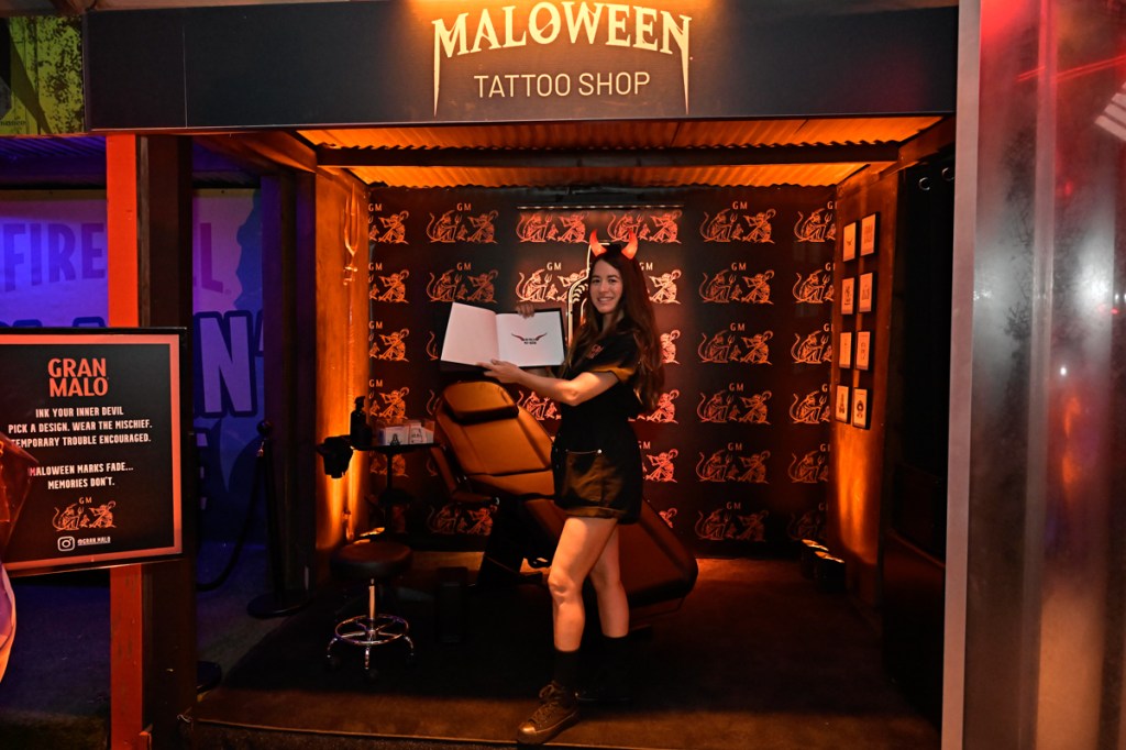 a girl wearing a black outfit and red devil horns holds up a book of tattoo designs inside a booth with a sign reading, "Malloween Tattoo Shop"