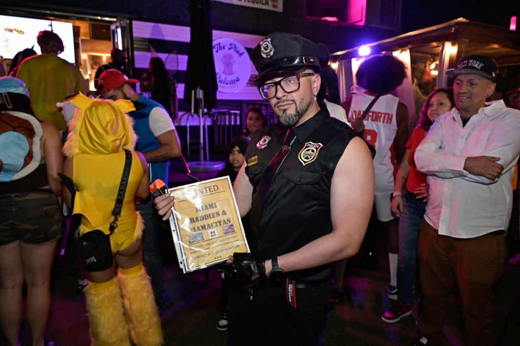 Photo of a man wearing a cop costume for Halloween. He holds a sign reading, "Wanted: Miami Baddies & Mamacitas"