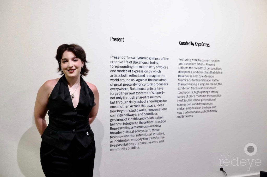 photo of a woman standing in front of an art gallery wall. The text on the wall indicates the exhibition is titled "Present," and is curated by Krys Ortega