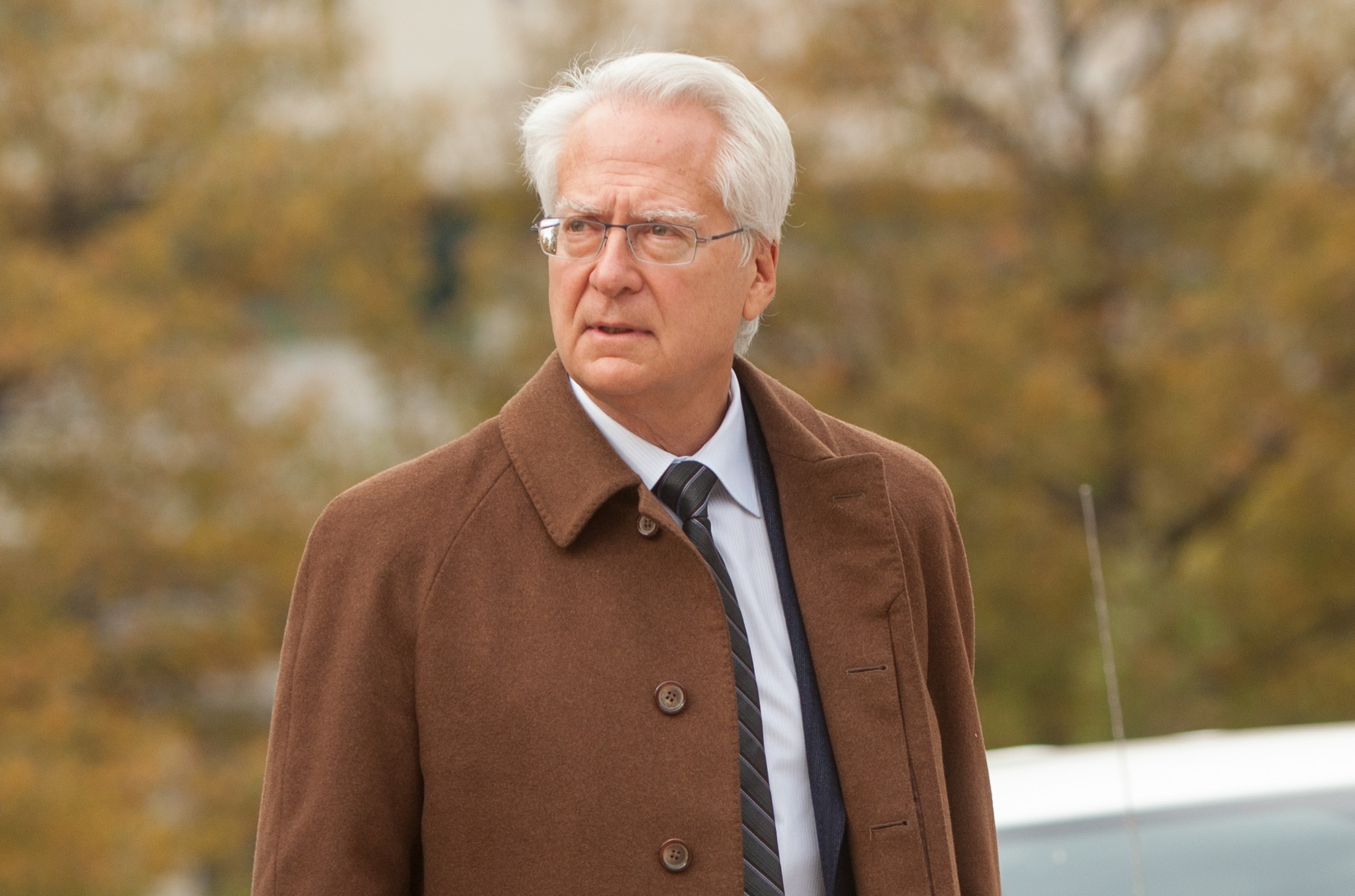 Photo of a bespectacled man with combed-back silver hair wearing a brown topcoat over a suit with blurred trees in the background. Shot in November in Maryland, as evidenced by fall colors.