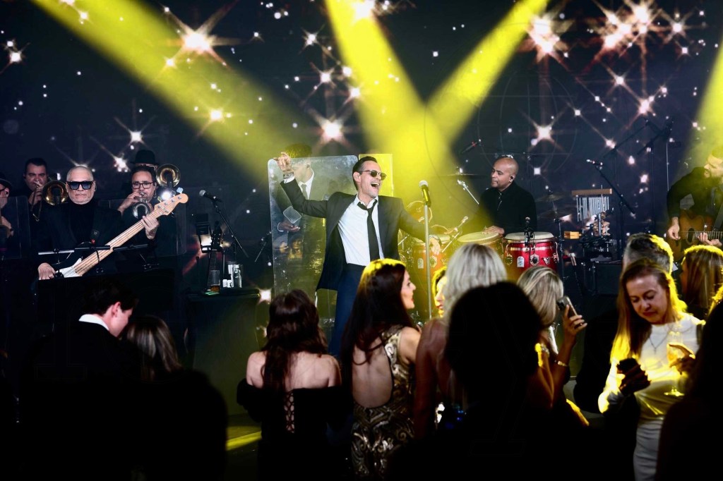 Photo of musician Marc Anthony performing onstage in a suit, backed by a bassist, conga player, and brass section. Yellow spotlights shine on him as he performs in front of an audience