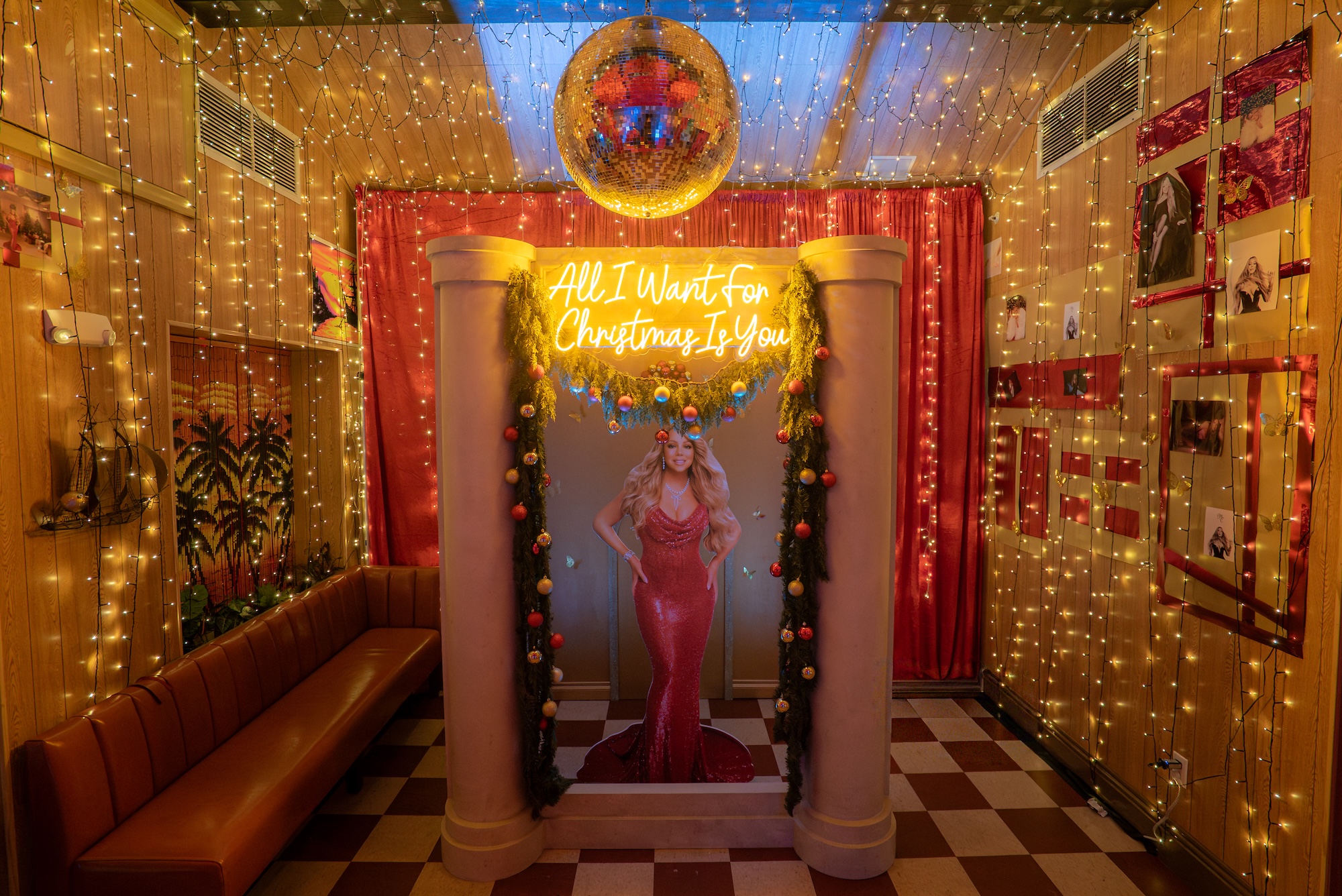 Photo of a bar with wood walls adorned in Christmas lights. In the center is a cardboard cutout of singer Mariah Carey wearing a tight, sequined red dress flanked by two columns. Above the display is a neon sign reading "All I Want for Christmas Is You"