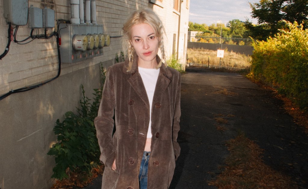 Photo of a blonde woman in pigtails, a long corduroy coat, and a white tee underneath, looking at the camera in an alley between a building and a hedge. Behind her is a field of weeds behind a fence