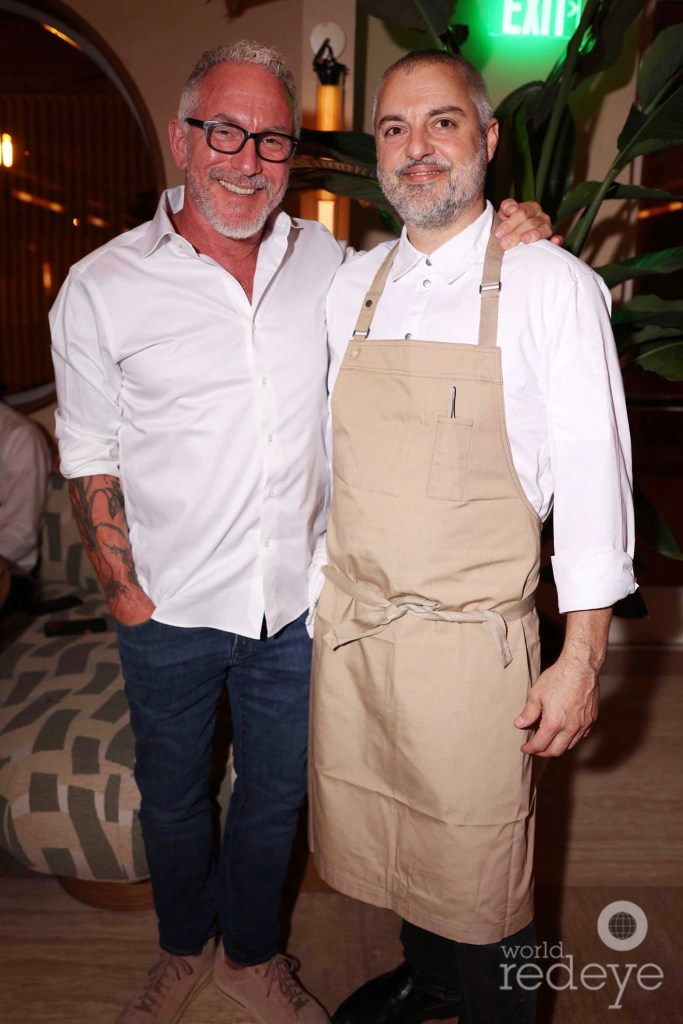 Photo of two men posing together. The man on the left wears jeans and a white button-down shirt. The man on the right wears a button-down long-sleeved shirt and a chef's apron.