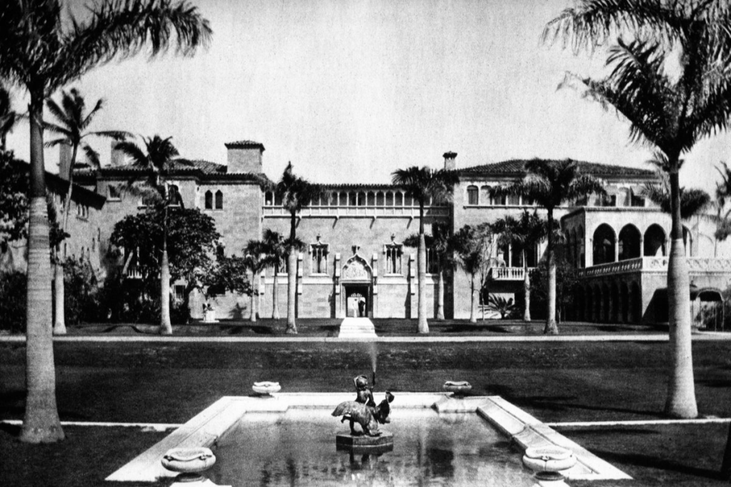 A massive lavish Spanish-style home sits behind an ornate water fountain.