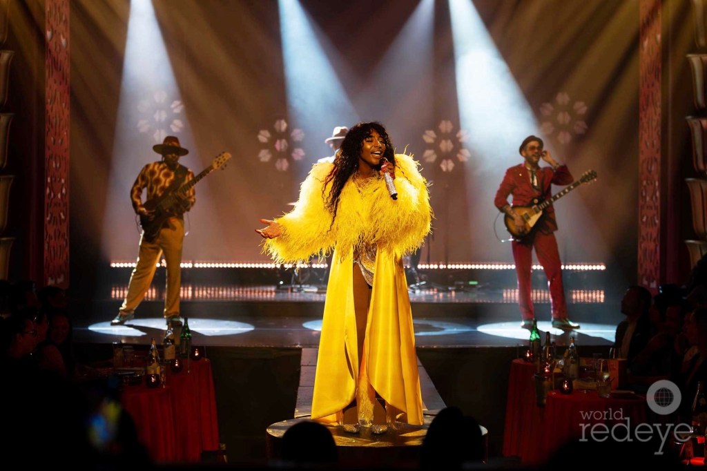 photo of a singer in a yellow dress and fuzzy coat performing onstage with three musicians behind her