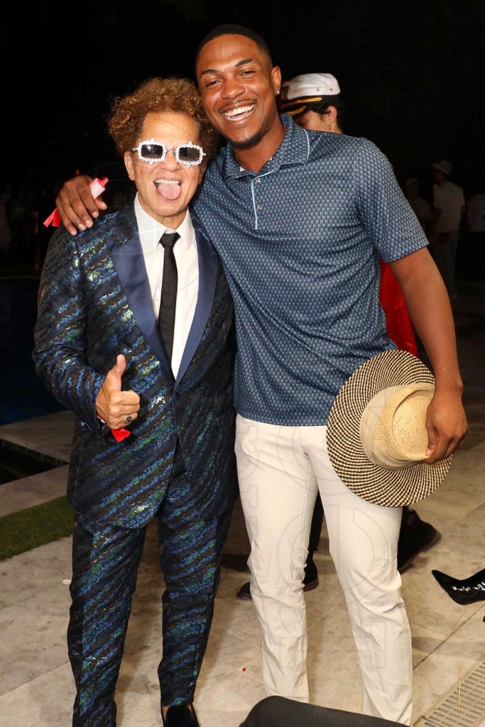 photo of a man in a scaled, iridescent suit and rhinestoned shades posing with his tonhue out next to a man in a jersey and slacks