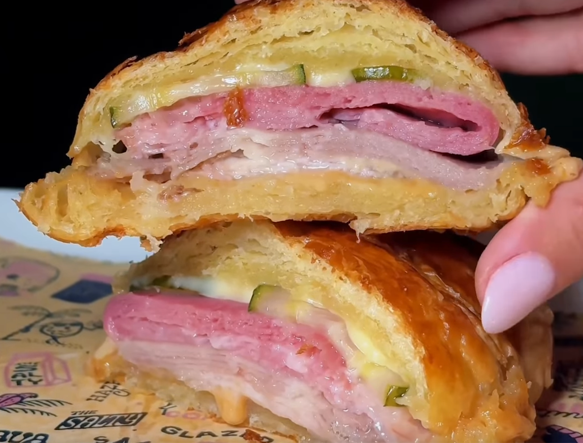a croissant filled with ham, cheese, and pickles next to a woman's finger