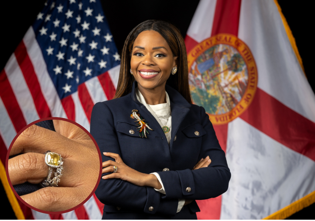 A Black woman stands with her arms folded in front of flags for the State of Florida and United States of America.