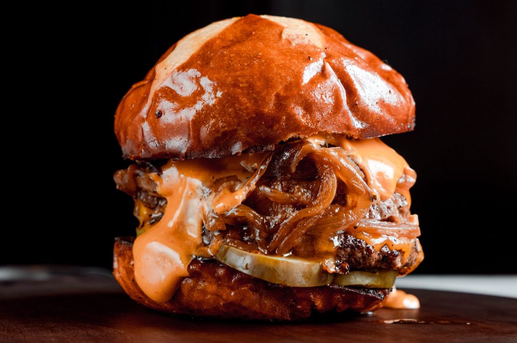 a loaded onion and cheeseburger on a pretzel bun with a black background
