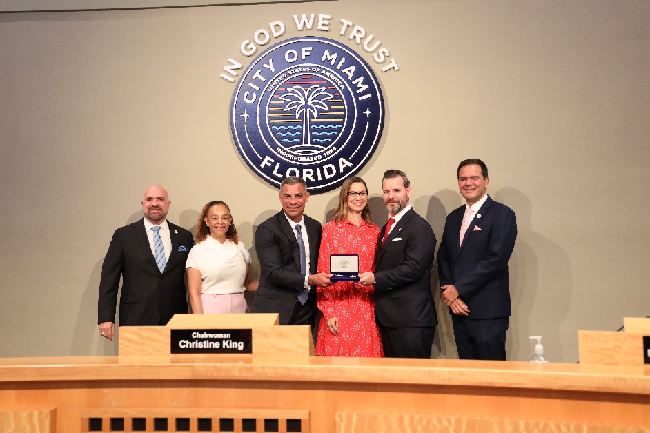 flanked by a group of people, a man poses with a smile while giving a box to another man in front of a City of Miami crest