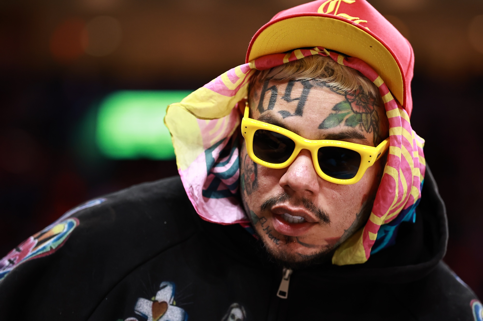 A rapper with multiple face tattoos wearing bright yellow-framed sunglasses and sporting colorful headgear courtside at an NBA basketball game