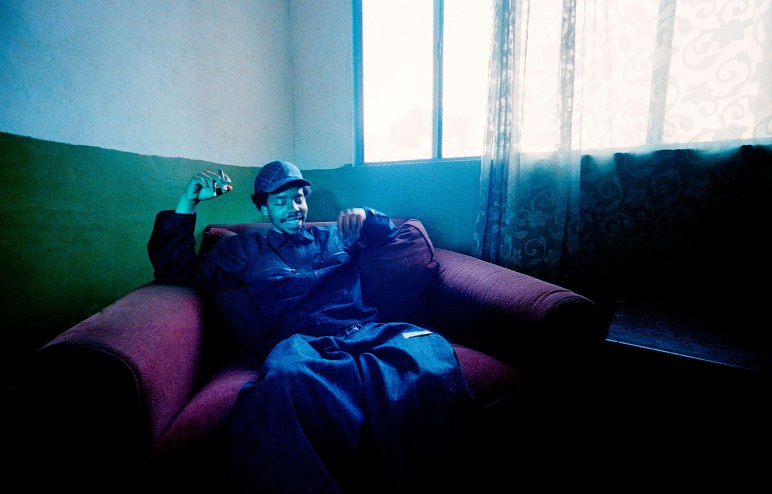 grainy photo of rapper Earl Sweatshirt lounging on a maroon plush chair in a room lit by natural light from an open curtain with a lace curtain on it. Sweatshirt wears a denim sweatshirt, baggy jeans, and a cap