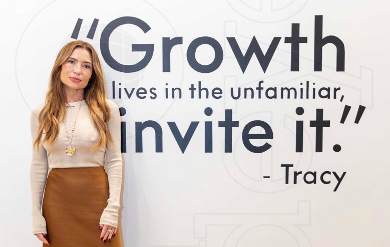photo of a woman in a beige long-sleeved shirt and brown skirt standing in front of a wall with writing reading, "Growth lives in the unfamiliar, invite it. -Tracy"