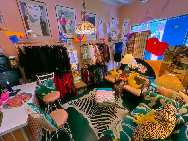 photo of a vintage shop with pastel-pink walls, teal-and-white couches, racks of clothing, and framed posters on the walls