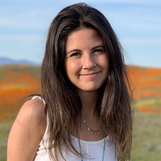 color photo of a smiling young woman with longish, dark hair, in a rural outdoor setting on a bright sunny day