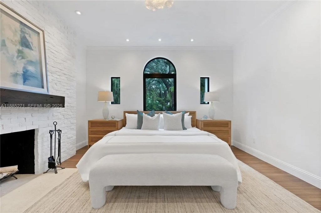 a bedroom is shown with a large bed in the center beside a fireplace capped by a black mantle holding a large painting