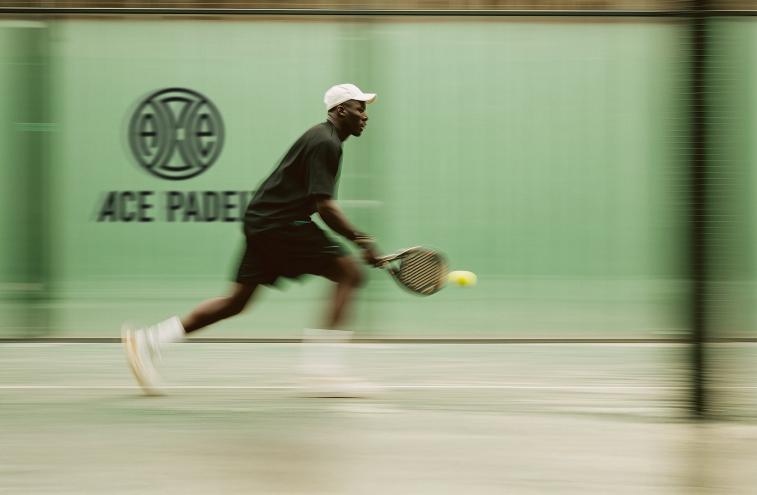Photo of a man playing padel inside a room with green walls. A sign on the wall reads, "Ace Padel"