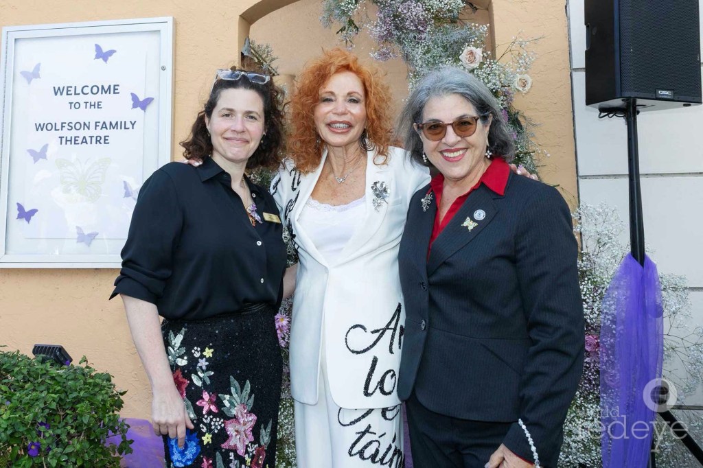 photo of three women posing to the right of a sign reading, "Welcome to the Wolfson Family Theatre"